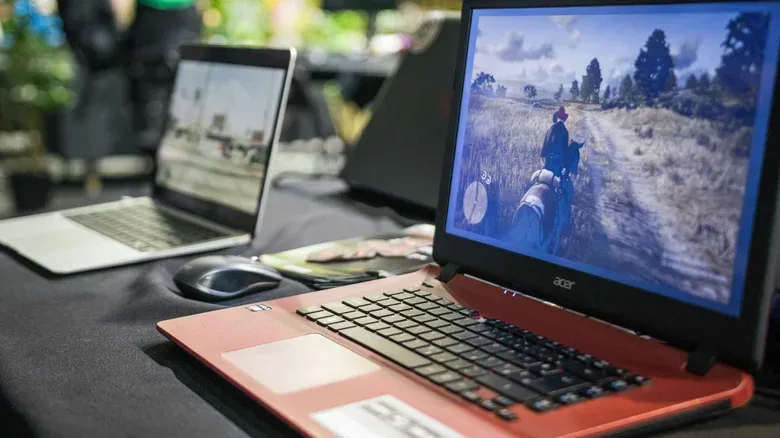 An old Acer laptop running Red Dead Redemption. Another laptop (blurred) is visible behind.