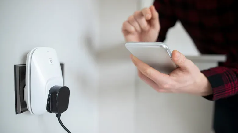 A person uses a smartphone beside a smart plug connected to a wall outlet.