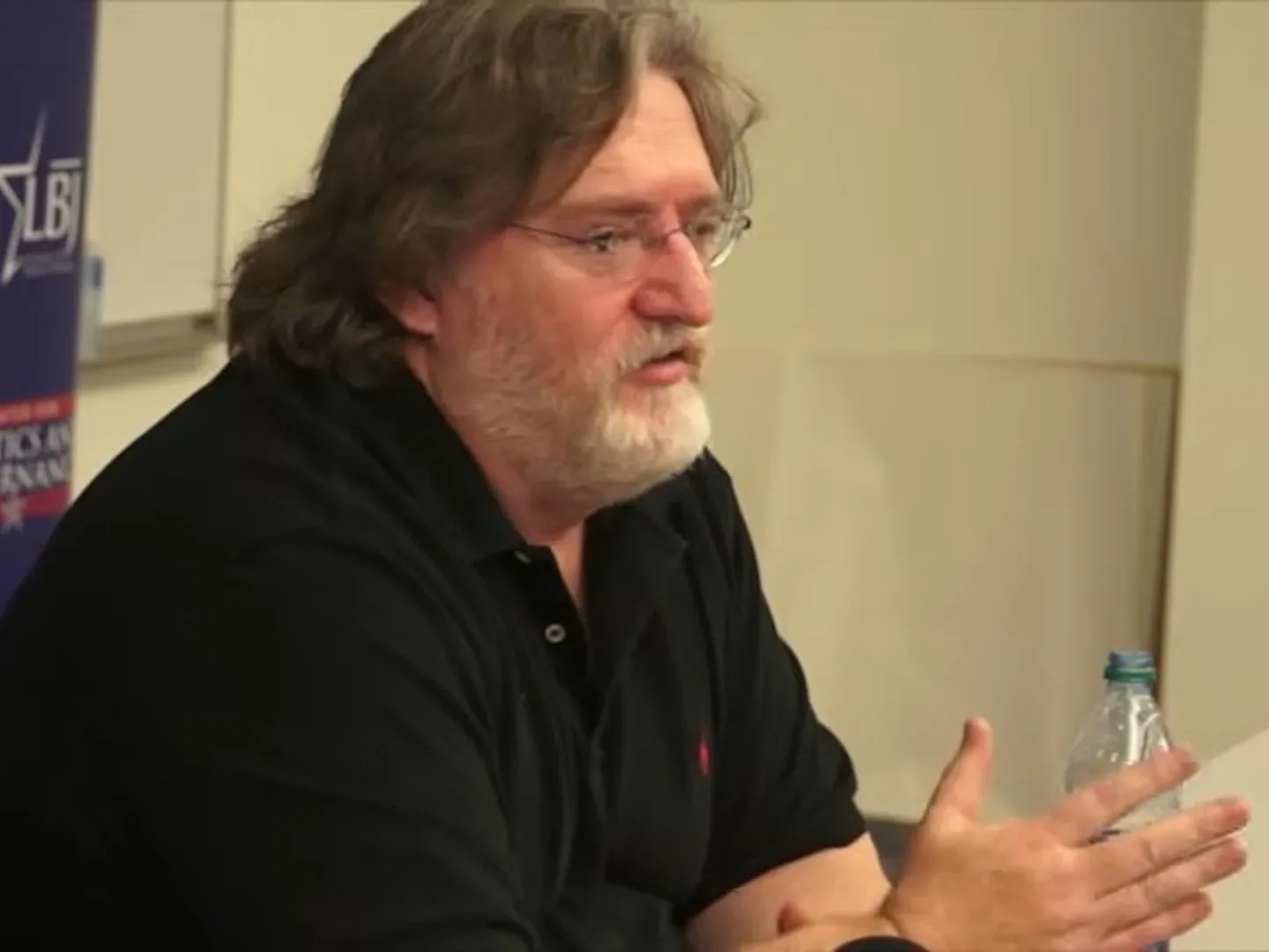 Gabe Newell in black shirt sitting at white desk for 2013 university talk