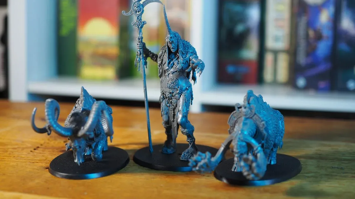 Ma Maegran and two monster models, all on a wooden surface in front of a board game shelf