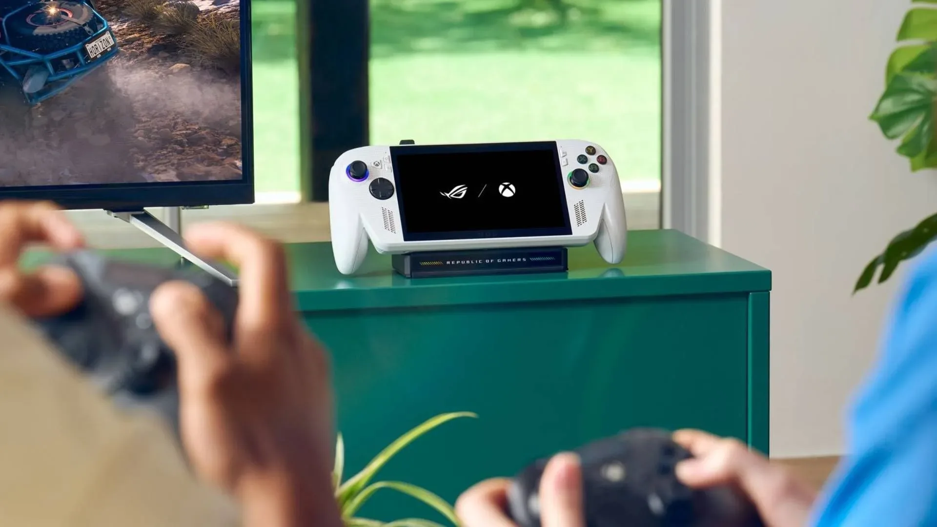 ROG Xbox Ally on green TV unit with hands in forefront holding controllers out of focus.