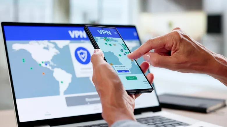 Person selecting a VPN server location on a smartphone while a laptop in the background displays a VPN map.