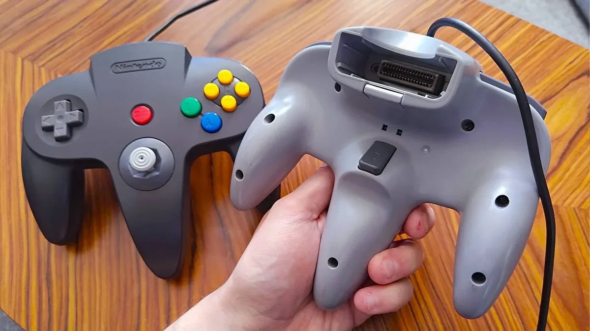 Hand holding gray N64 controller with Z button facing forward and black N64 gamepad on table in backdrop.