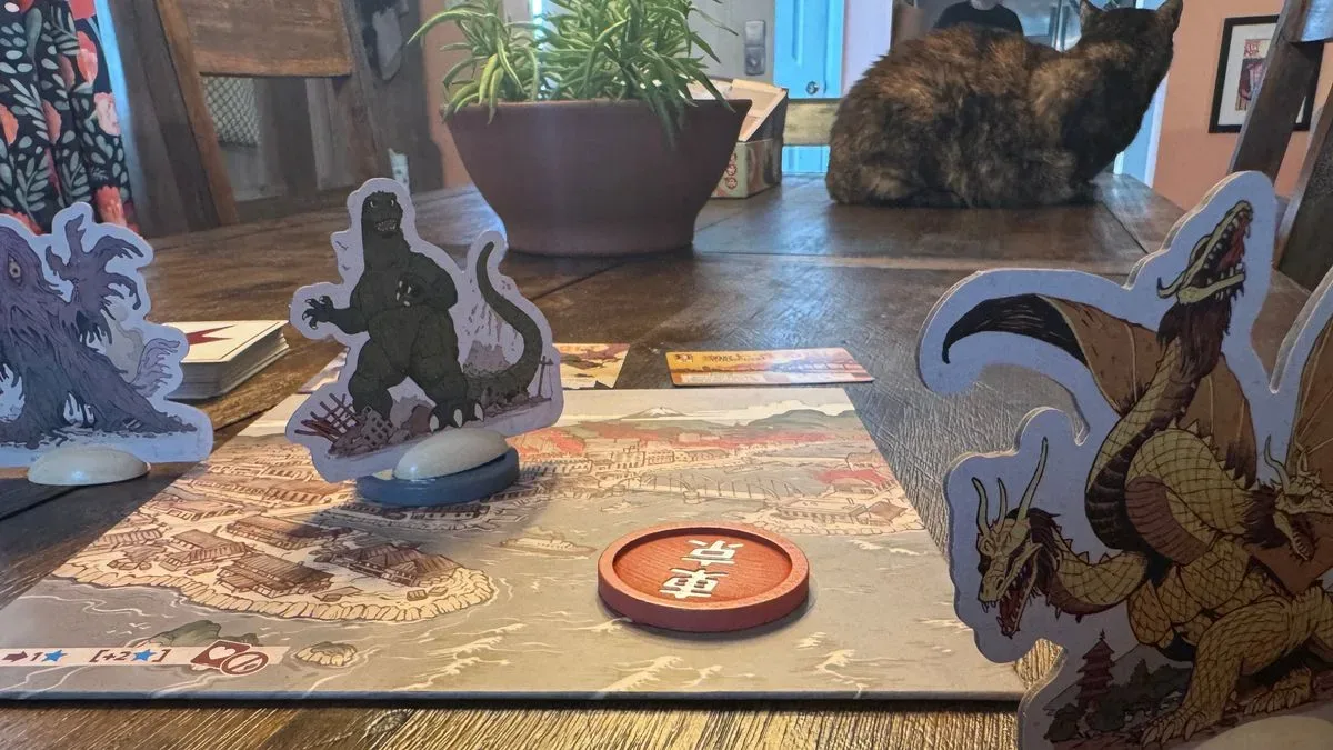 Multiple King of Tokyo: Godzilla tokens and cards on a wooden surface, with a cat visible in the background