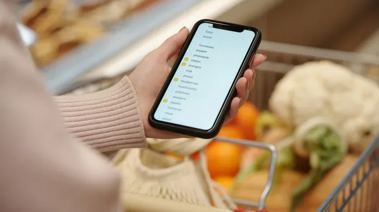 A shopper checks a smartphone while pushing a grocery cart through a produce aisle.