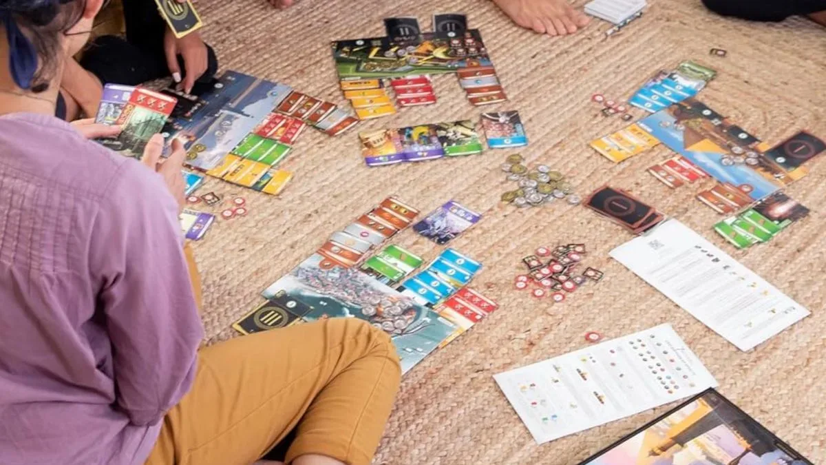 A host of players gather around the boards, cards, and tokens of 7 Wonders as it's played on the floor