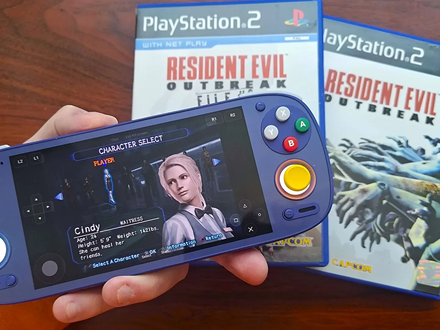 Hand holding Retroid Pocket 5 with Resident Evil Outbreak web version running and Cindy selected on character screen with original PS2 copies of File #1 and File #2 in backdrop.