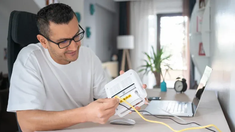 Man hooking up Ethernet cables to a router on a desk, next to a laptop.