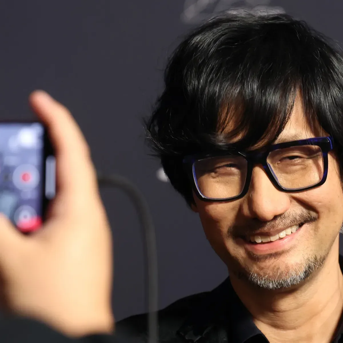 Hideo Kojima standing outside Sony City in Tokyo wearing a Joy Division T-shirt