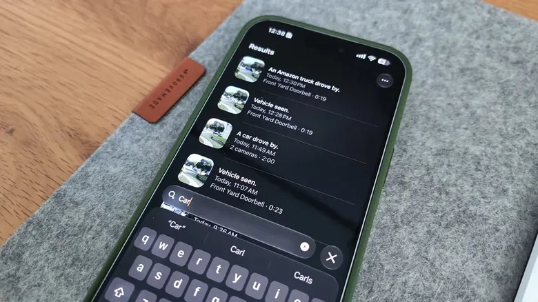 A green iPhone sits on a grey folder atop a wooden desk, where a user types Car into the Home app's search engine. A lit of videos where a car drove by appear on screen.