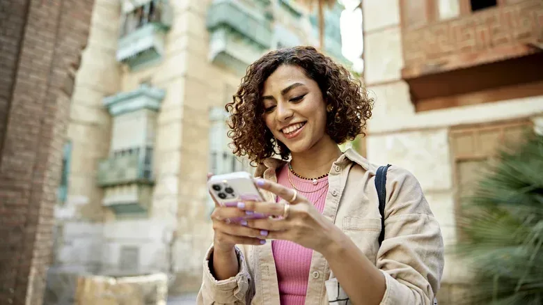 A woman in a pink shirt and tan jacket uses her phone as she walks away from a brick apartment building