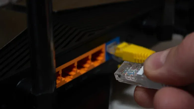 A hand holding a clear Ethernet connector beside a black router with orange LAN ports and a yellow cable connected to a blue port.